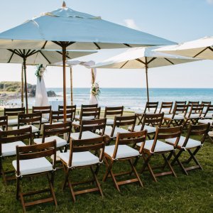 Rows of wooden chairs with white cushions arranged under large white umbrellas face an ocean view at an outdoor event setup on grass.