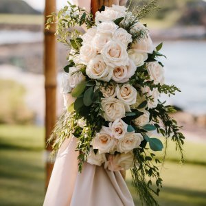 A floral arrangement of white roses and greenery decorates a wooden arch with a scenic outdoor background.