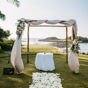 A wooden arch draped with white fabric and floral arrangements stands by the ocean. A round table covered with a white tablecloth is beneath the arch, and a white petal aisle leads to the setup.