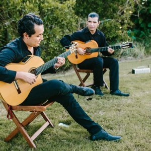 Two men in black attire play acoustic guitars while sitting on wooden chairs outdoors on the grass, surrounded by greenery.