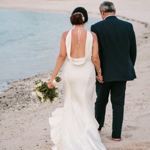 A couple dressed in formal wedding attire walks hand in hand along a beach, with the bride holding a bouquet and the ocean in the background.