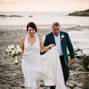 A bride holding a bouquet and lifting her dress hem walks alongside a man in a suit on a rocky beach at sunset.