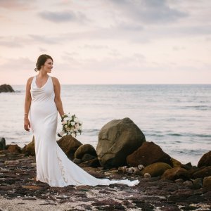 A woman in a white dress holding a bouquet stands on a rocky beach with the ocean and a cloudy sky in the background.