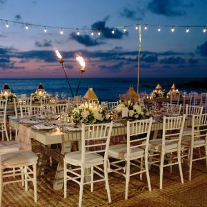 A beachside wedding reception setup at sunset with white chairs, floral centerpieces, and string lights overhead.