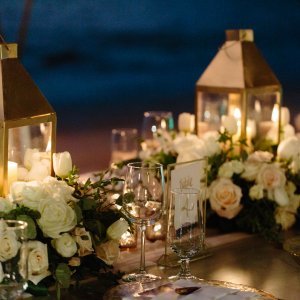 A close-up of an elegantly set table with floral arrangements, candles in lanterns, and glassware, designed for an evening event.