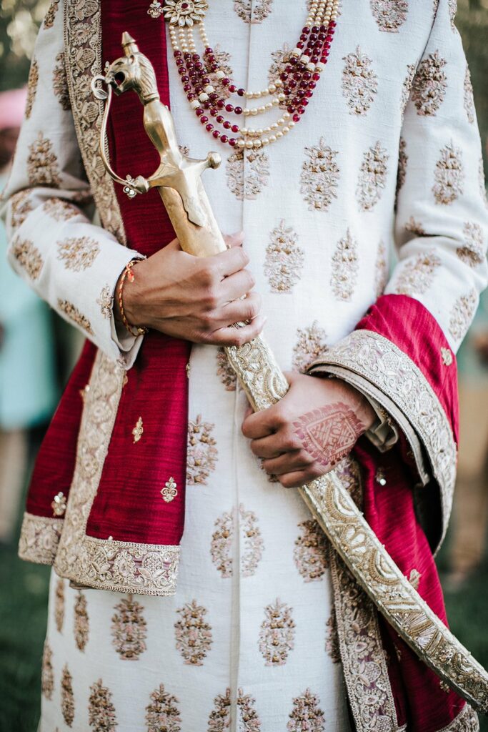 A person dressed in ornate traditional attire holds a decorative sword. The outfit includes a white and gold embroidered garment with red accents and layered necklaces.