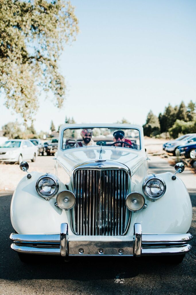 Two people driving a vintage white convertible car on a sunny day, with trees and parked cars in the background.