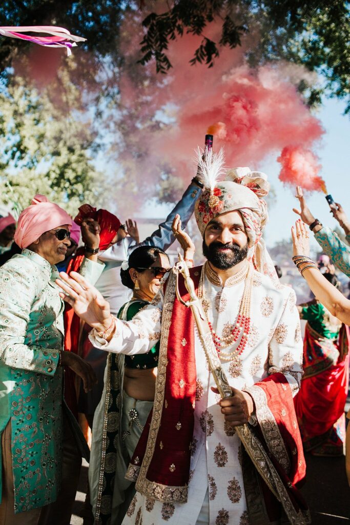 A person in traditional attire, holding a staff, leads a procession outdoors. Colorful smoke and festive clothing are visible in the background.