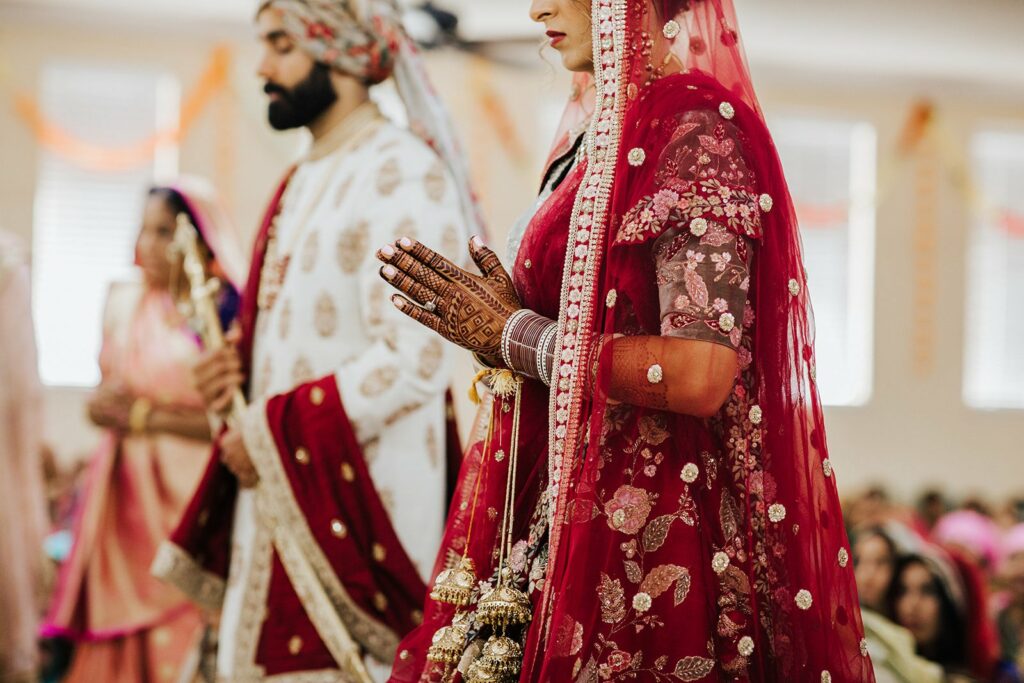 A couple dressed in traditional Indian wedding attire stands with hands clasped in what appears to be a ceremonial setting. The bride wears a red embellished outfit; the groom is in white and gold.
