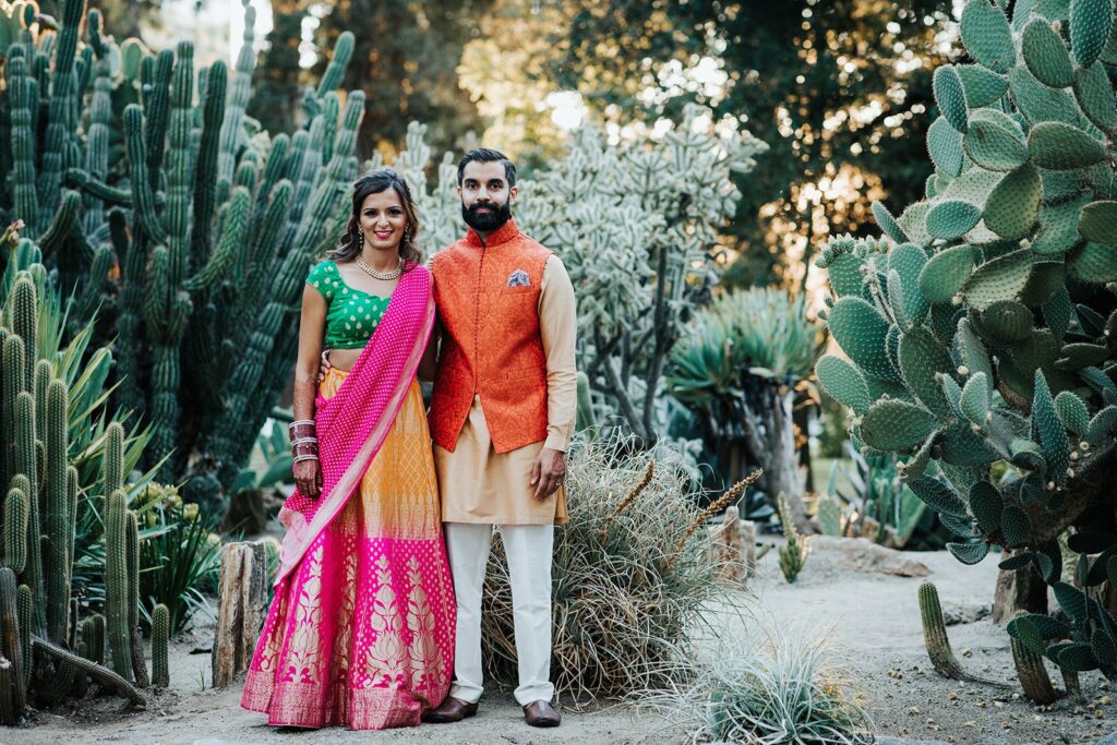 A man and woman in traditional attire stand in a garden surrounded by various cacti and succulents, smiling at the camera.