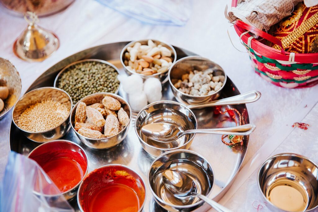 A stainless steel tray holds various spices, seeds, nuts, and small bowls with spoons. A woven basket with cloth and other items is nearby on a white surface.