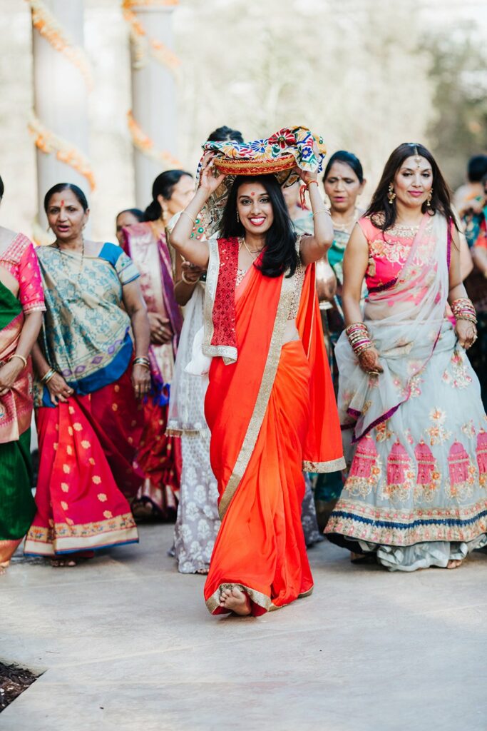 A woman in a bright red sari carries a colorful basket on her head while walking with a group of women in traditional attire.