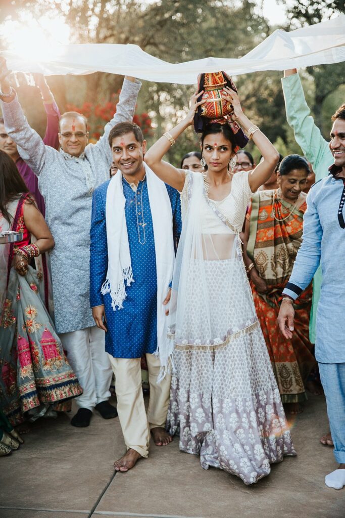 A bride in traditional attire holds a decorated pot on her head while guests surround and support her with a cloth overhead during a wedding ceremony.