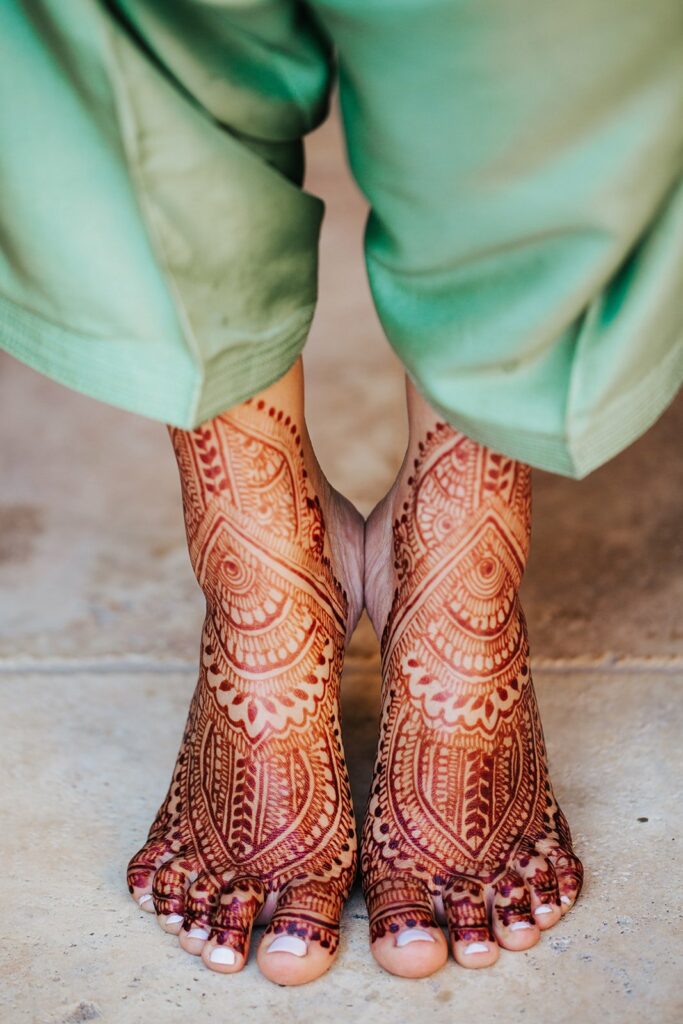 Close-up of feet with intricate henna designs extending from the toes to the ankles. The person is standing, and their green pants are visible.