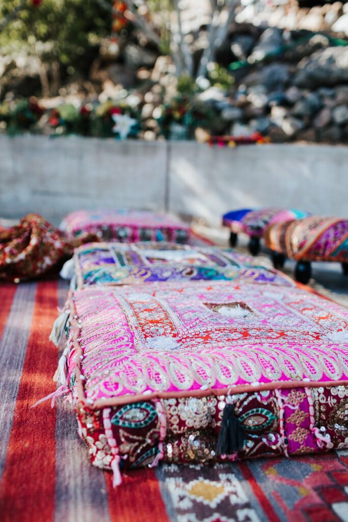 Vibrant, intricately patterned cushions and rugs are laid out on a surface outdoors, with a stone wall and foliage visible in the background.