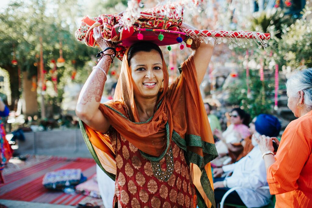 A woman in traditional attire carries a decorated object on her head during an outdoor event. Others in traditional clothing are in the background.