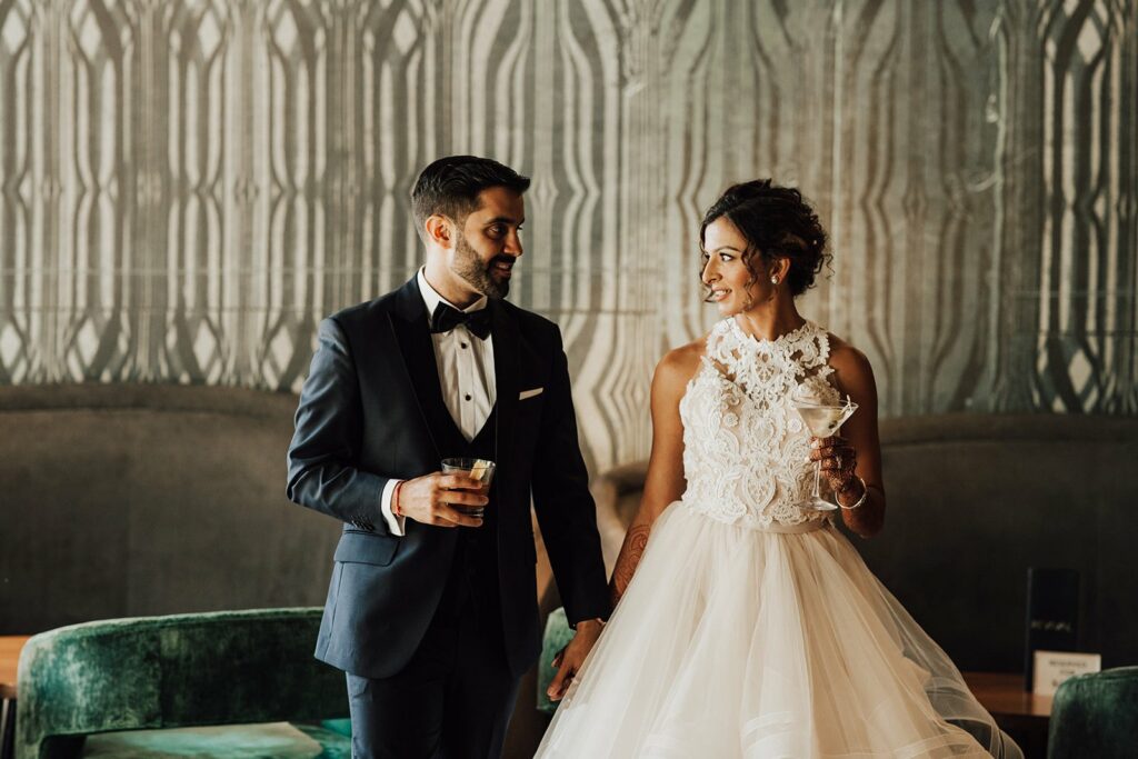 A bride in a white gown and a groom in a black tuxedo hold hands and drinks while standing in an elaborately decorated room.