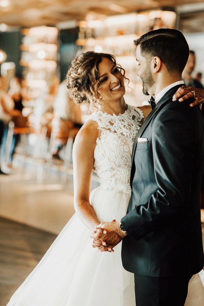 A couple dressed in wedding attire smiles at each other while dancing. The bride wears a white lace gown, and the groom is in a black suit.