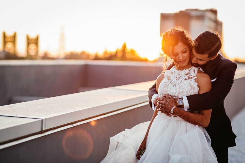 A man in a suit embraces a woman in a white gown on a rooftop at sunset, with buildings and trees in the background.