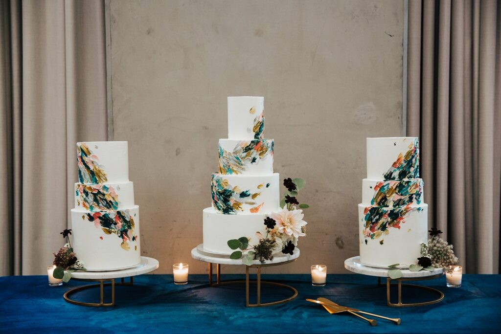 A display of three white tiered cakes with colorful abstract decorations on a blue tablecloth, surrounded by candles and flowers.