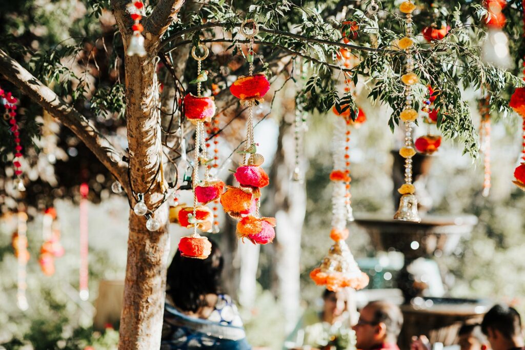 Colorful decorations hang from tree branches in an outdoor setting, with orange, pink, and yellow flowers and ornaments creating a festive atmosphere. People mingle in the background.