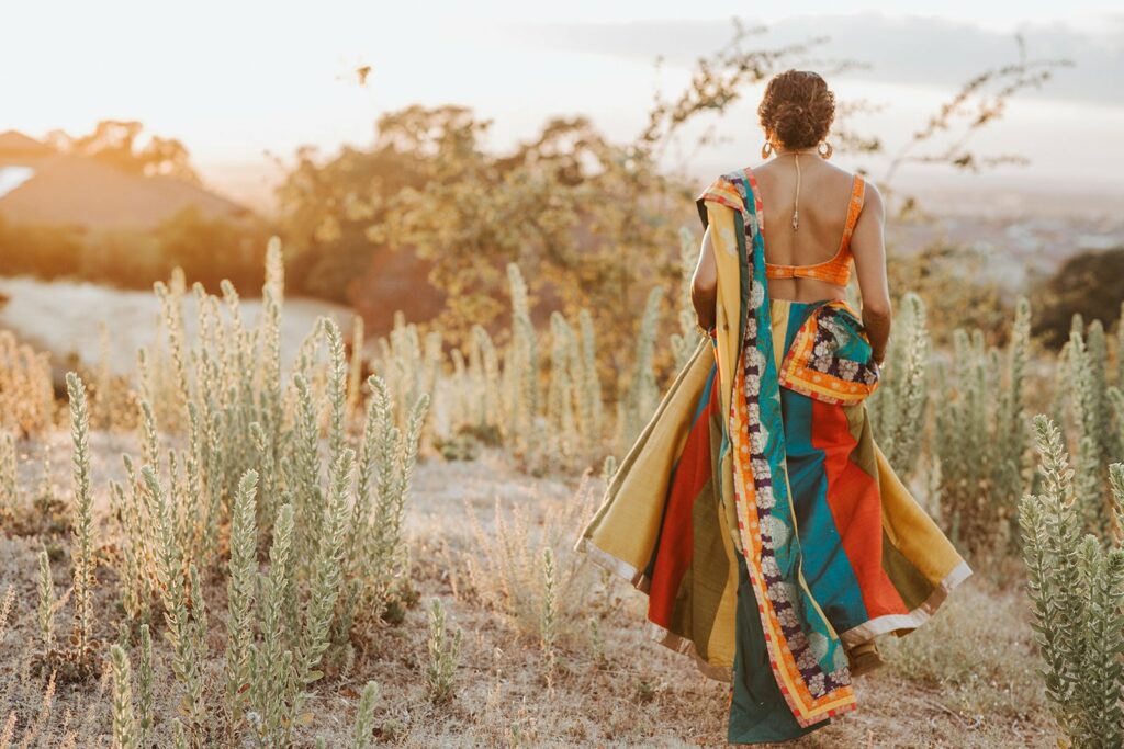 A person in traditional colorful attire walks through a field of tall grass and plants at sunset, with a view of the landscape in the background.