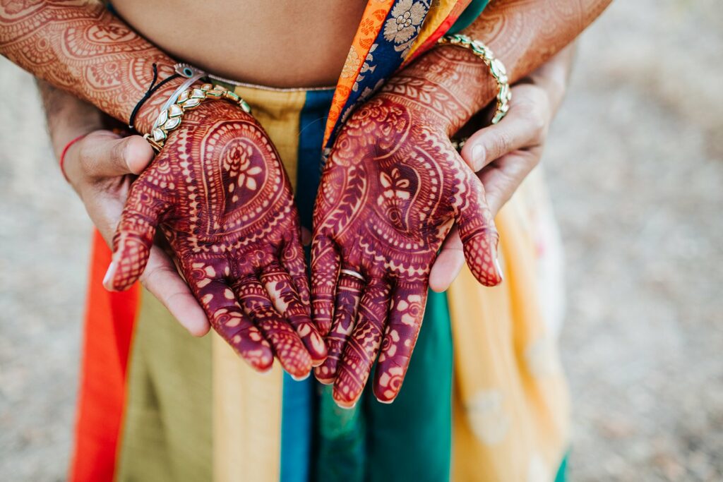 Two hands with intricate henna designs are cupped together, with a colorful, patterned garment visible in the background.