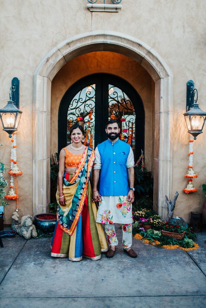 A woman and a man in colorful traditional South Asian attire stand in front of an ornate doorway decorated with flowers. They are holding hands and smiling.