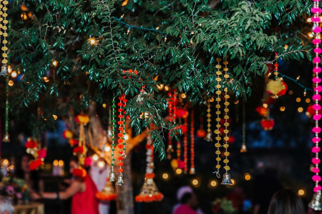 Outdoor scene with tree branches decorated with colorful hanging ornaments, including bells and lights. Blurred people in the background suggest a festive gathering or celebration at night.