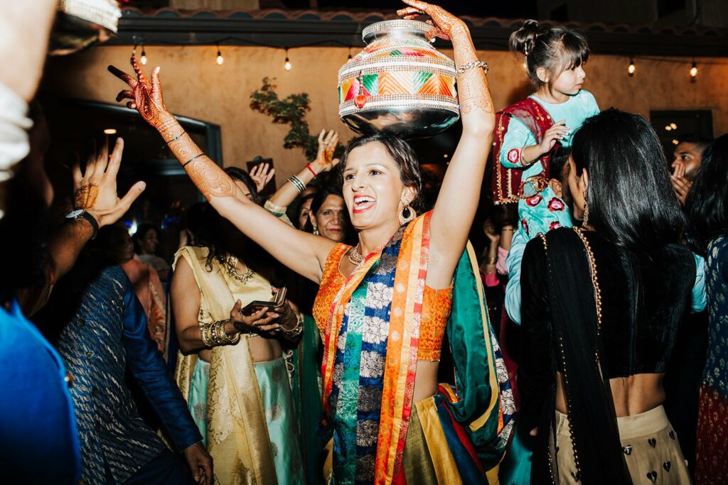 A woman in colorful traditional clothing dances joyfully, balancing a decorated pot on her head, surrounded by people at a lively celebration.
