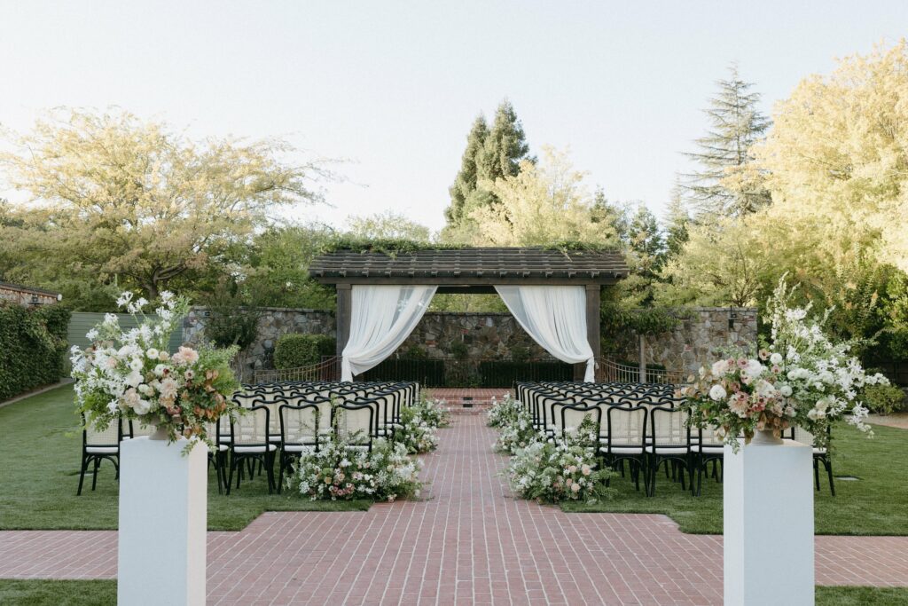 An outdoor wedding ceremony setup with two rows of black chairs facing an altar. The altar is under a wooden pergola with white drapes. Flower arrangements decorate the aisle and surroundings, meticulously arranged by Napa Wedding Planners for a perfect day.