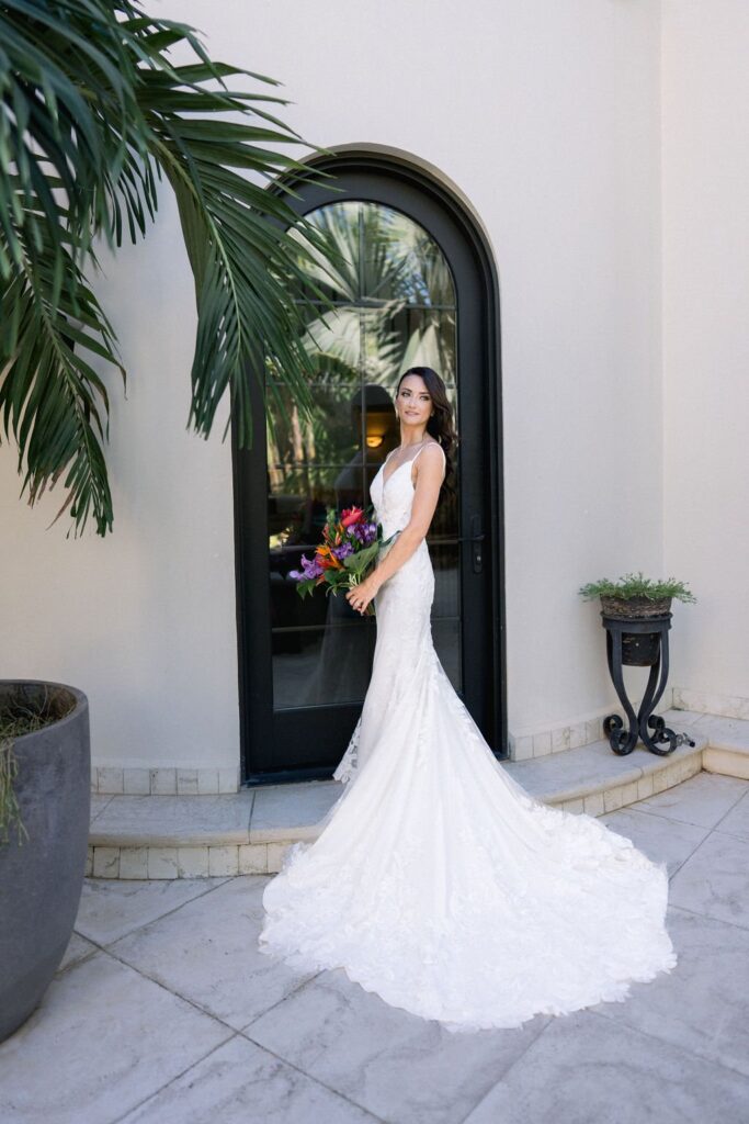 A woman in a white wedding dress holds a colorful bouquet and poses beside a black arched door, with green plants in the background.