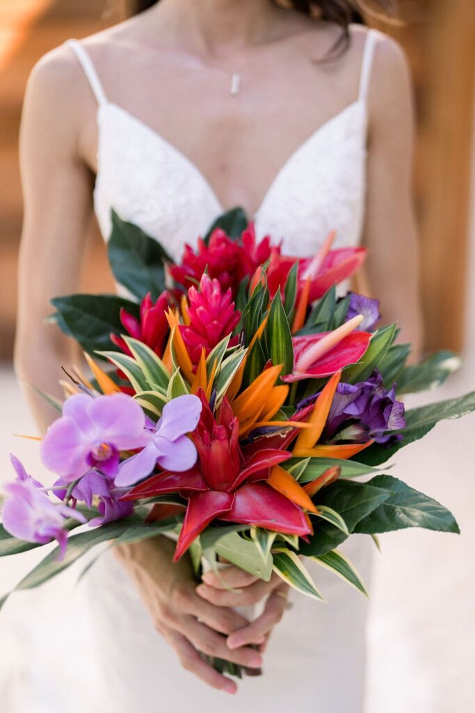 A bride in a white dress holds a vibrant bouquet of tropical flowers, featuring red, orange, pink, and purple blooms with lush green leaves.