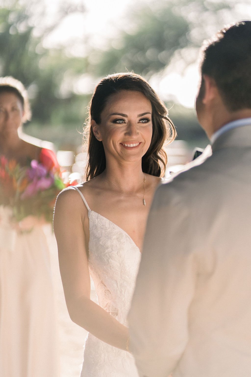 A bride smiles at her partner during an outdoor wedding ceremony, with a bridesmaid holding flowers in the background.