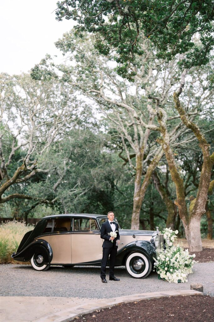 A person in formal attire stands beside a vintage car adorned with white flowers, surrounded by trees.