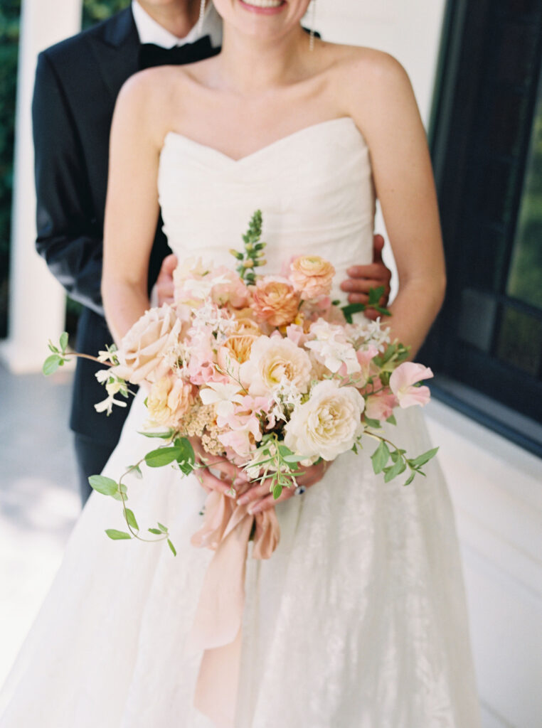 Bride holding a bouquet of white, pink, and peach flowers. Groom partially visible in the background.
