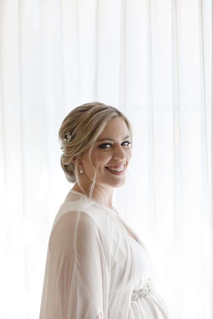 A smiling woman in a bridal gown stands in front of sheer curtains, looking over her shoulder.