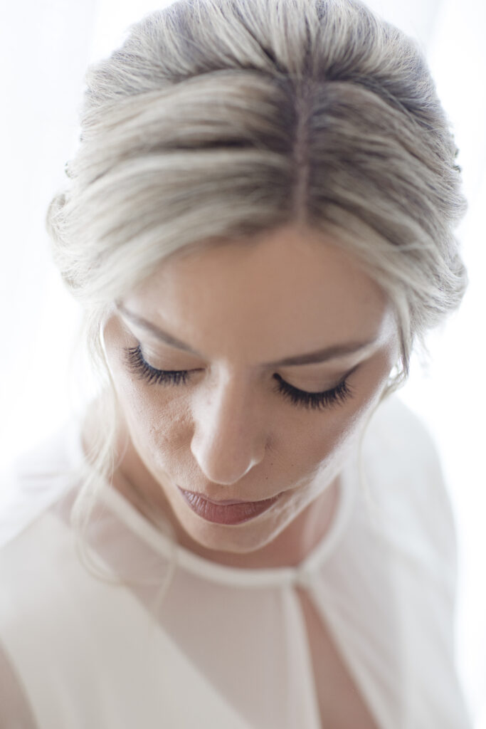 A woman with blonde hair styled up is looking downward. She has long eyelashes and is wearing a white blouse.