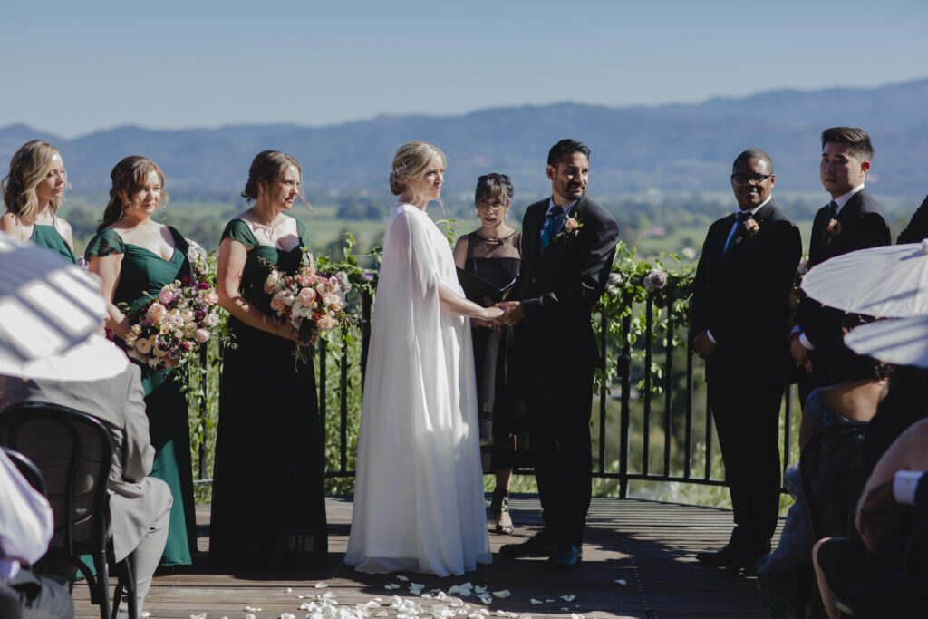 Bride and groom holding hands in an outdoor wedding ceremony with bridesmaids and groomsmen standing on either side.