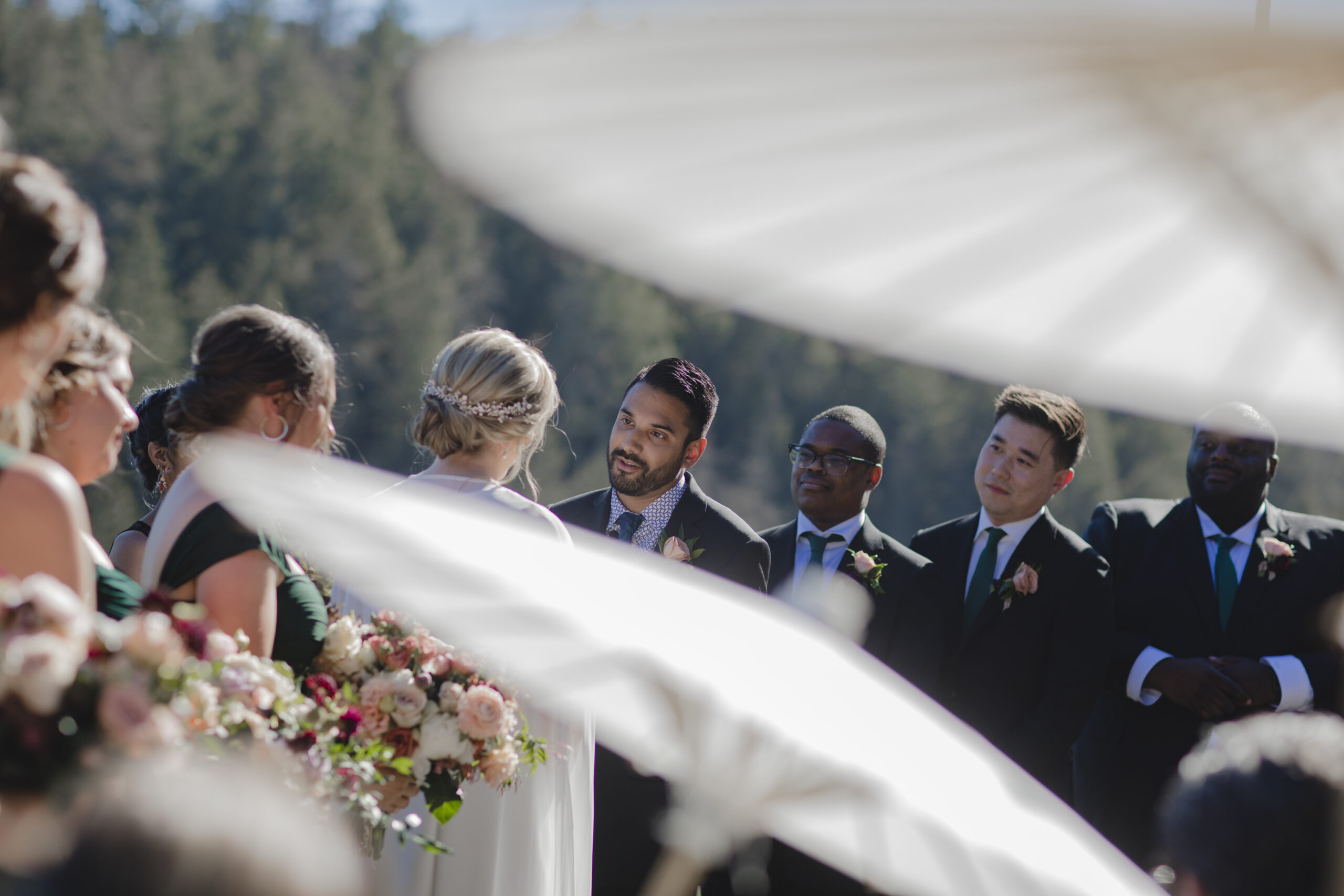 A wedding ceremony with a bride and groom standing before each other, surrounded by guests and groomsmen. Bouquets and white parasols are partially visible in the foreground.