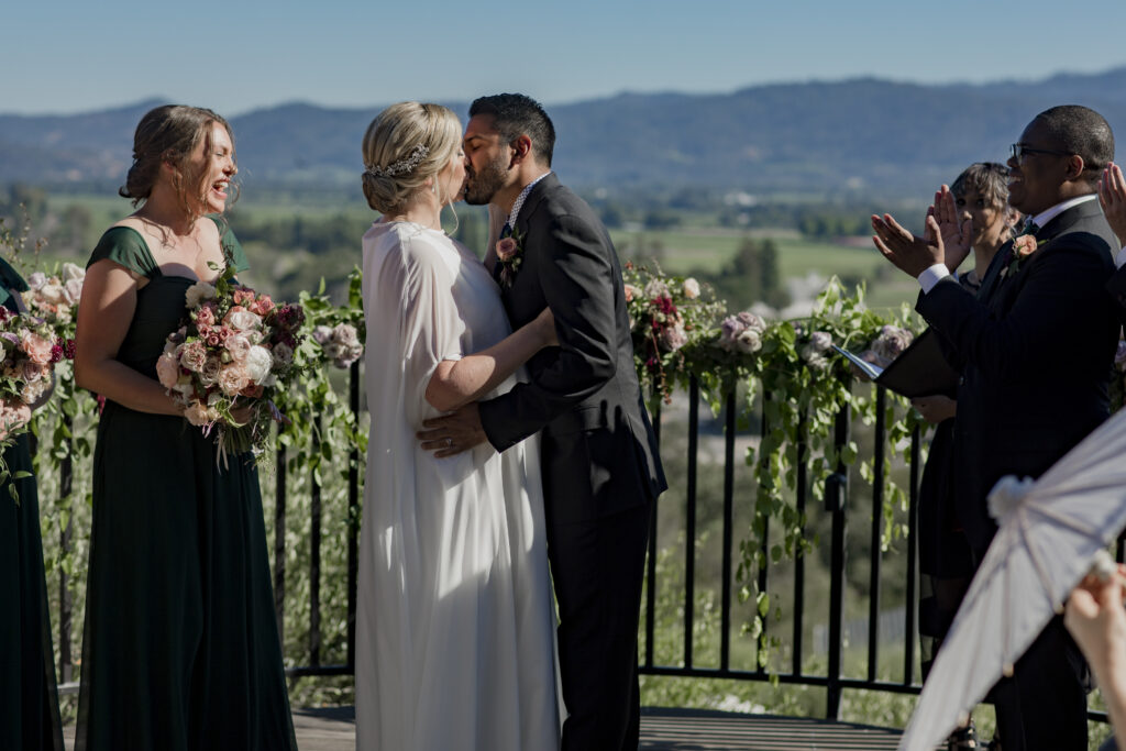 A wedding couple kisses at an outdoor ceremony. Two bridesmaids stand beside them holding bouquets, and an officiant claps in the background. Greenery and flowers decorate the railing.