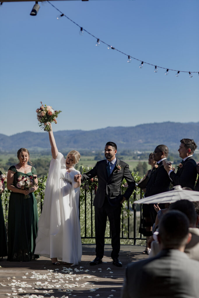 Bride and groom stand on an outdoor deck; the bride raises her bouquet. Bridesmaid and groomsmen are nearby. Mountains and a clear sky form the background.