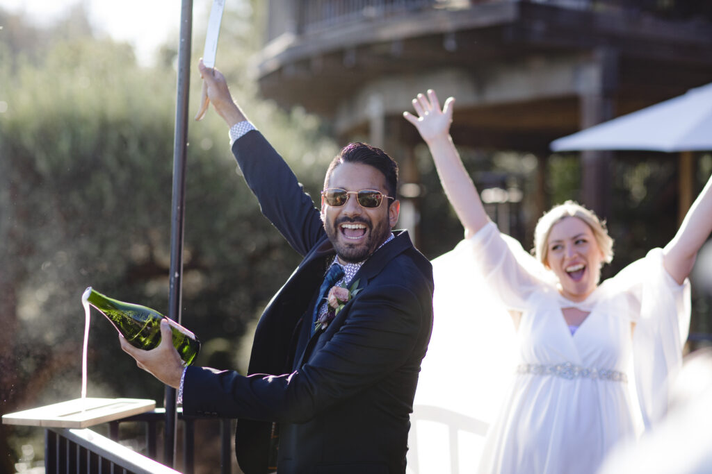 A man in a suit joyfully opens a bottle of champagne while a woman in a white dress raises her arms in celebration outdoors.