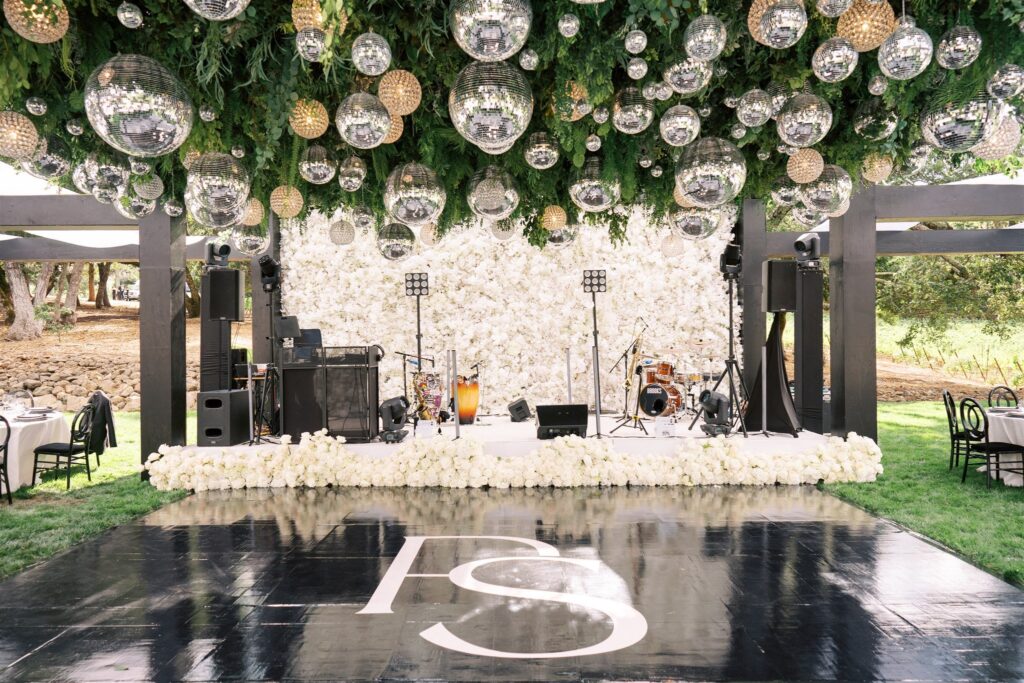 Elegant outdoor stage with white floral backdrop, musical instruments, and reflective spheres hanging from greenery above a glossy black floor with initials "PS.
