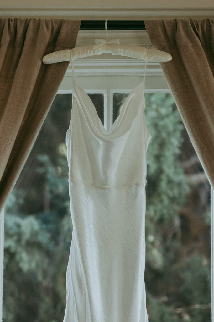 A white satin slip dress hangs on a padded hanger in front of a window with brown curtains, greenery visible outside—a serene scene reminiscent of a Fall Park Winters wedding.