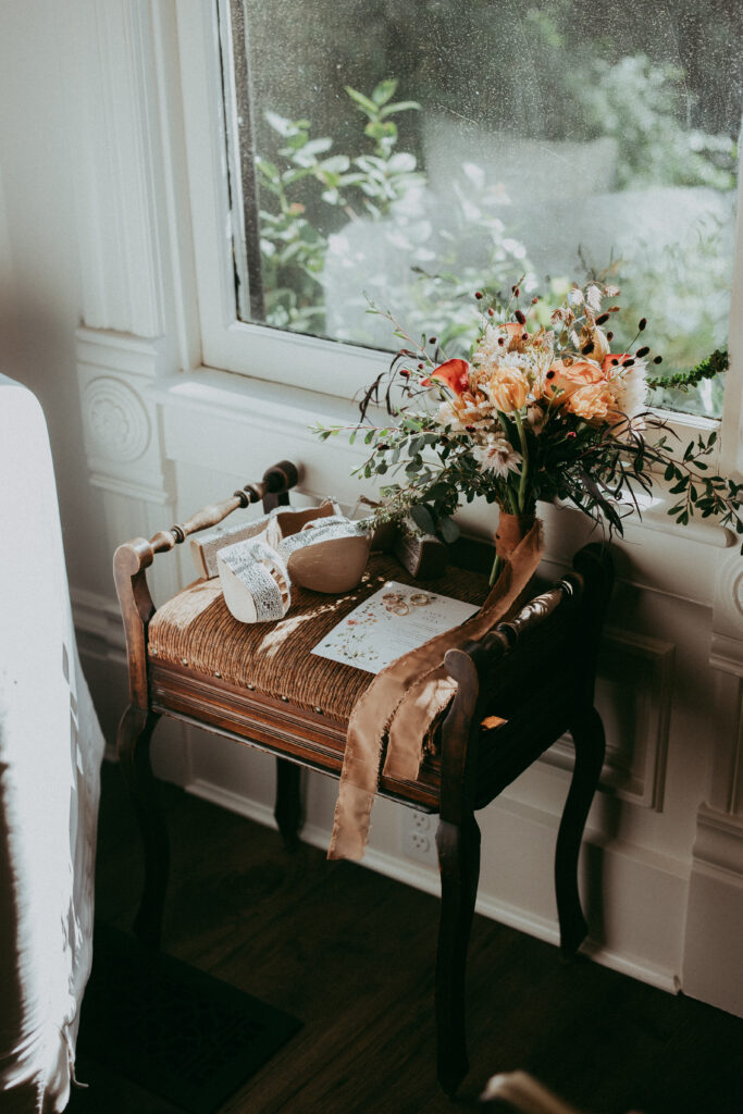 A small wooden bench holds a bouquet of flowers in a vase, a ribbon, a ceramic bowl, and a decorated box, placed by a window with sunlight streaming in—capturing the cozy charm of a fall wedding.