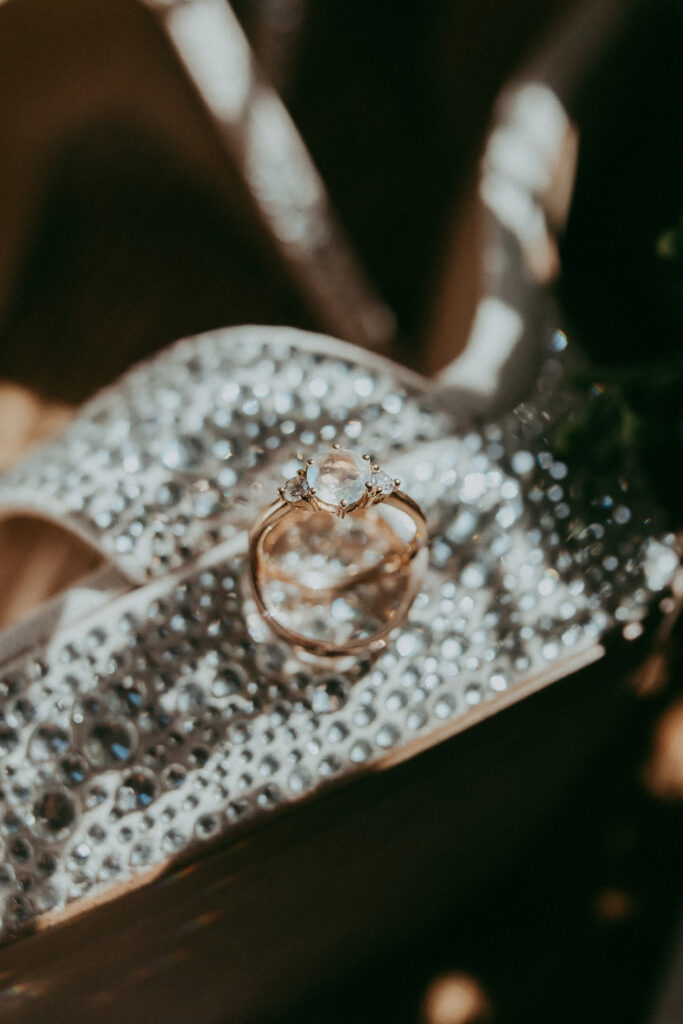 Two gold rings, one with a clear gemstone, rest on a sparkling, rhinestone-encrusted surface, capturing the elegance of an autumn celebration—perfect for a Fall park wedding.