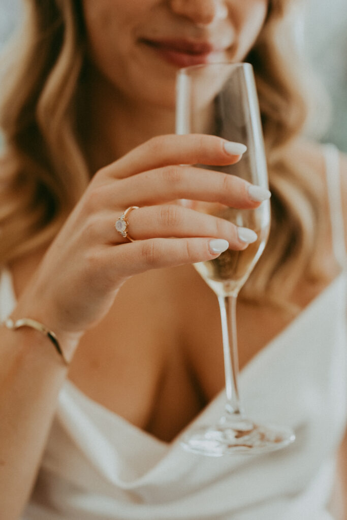 Woman in a white dress holding a champagne glass, showing a diamond ring on her finger, with light pink nail polish and wavy blonde hair, captured during a romantic fall park wedding.