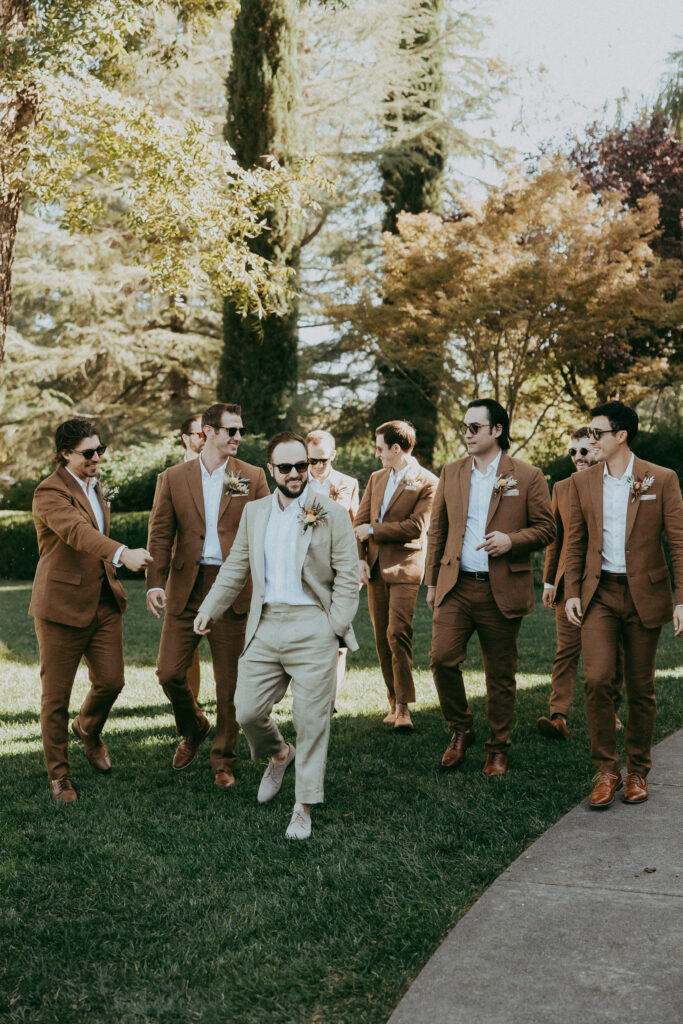 A groom in a light suit walks with seven groomsmen in matching brown suits, all outdoors on a sunny day at a picturesque park, surrounded by trees and grass—a perfect scene for a classic fall wedding.