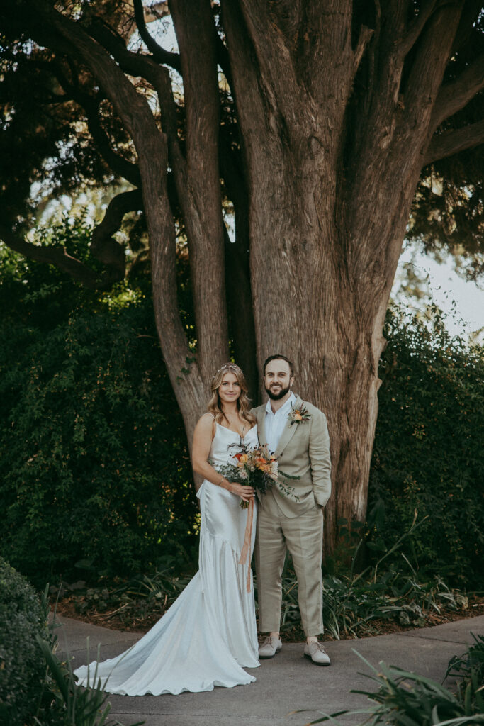 A couple in wedding attire stands together outdoors in front of a large tree in a beautiful park, with lush greenery and hints of fall colors setting the perfect scene for their memorable wedding.